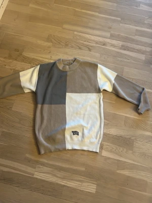 Unik Burberry Sweatshirt - Säljer en Burberry stickad tröja i beige, grå och vit blockdesign. Tröjan har rund hals, långärmade och ett broderat Burberry-logomärke framtill. Ribbad struktur och klassisk passform, perfekt för dig som gillar stilrena och neutrala färger.