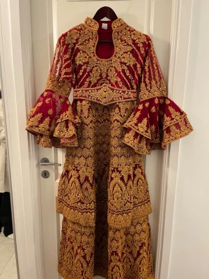 Vinröd ”Henna” långklänning med guld brodyr - Superfin vinröd långklänning med lyxig guld brodyr och detaljer. Klänningen har vida volangärmar, hög krage och markerad midja. Perfekt för bal eller fest. Materialet är troligtvis polyester eller sammet med broderade detaljer.