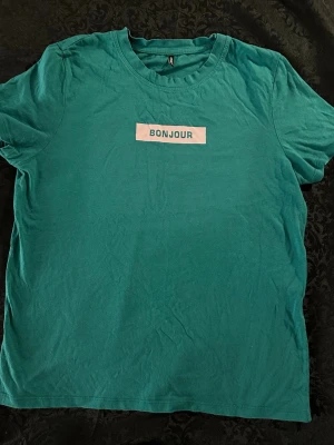 Grön t-shirt med BONJOUR-tryck storlek M  - T-shirt i klargrön färg från ONLY med vitt tryck där det står 'BONJOUR' på bröstet. Klassisk rund halsringning och korta ärmar. Tillverkad i mjuk bomull som känns skön mot huden. Perfekt för dig som gillar enkel men snygg stil.