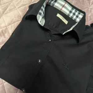 Svart skjorta från Burberry - Snygg svart skjorta från Burberry med klassisk mönster i kragen och armarna. Den har en diskret broderad logga på bröstet. Skick 8-10 passar XS/XXS kan gå ned i pris vid snabb affär. 