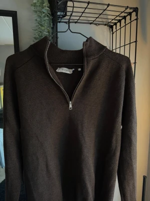 Brun stickad tröja med half zip SANDÖ - Mörkbrun stickad tröja från SANDÖ i storlek M. Tröjan har en hög krage och en snygg half zip-dragkedja framtill. Perfekt för lager på lager och passar till både jeans och chinos. Långärmad och mjuk i materialet.