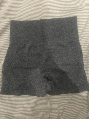 Grå seamless träningsshorts - Säljer ett par grå seamless träningsshorts med hög midja och scrunch-detalj bak. Materialet är stretchigt och sitter tight mot kroppen, perfekt för gymmet eller andra sportaktiviteter. Shortsen har ribbade sidor och meshpaneler för extra ventilation.