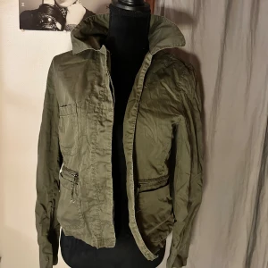 Olivgrön vindjacka med dragkedjor - Snygg olivgrön vindjacka med hög krage och praktiska fickor med dragkedjor. Jackan har en enkel design med långa ärmar och är tillverkad i ett lätt bomullsmaterial. Perfekt för dig som gillar en chill och urban stil.