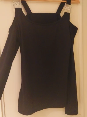Svart långärmad off sholders tröja  - Säljer en svart långärmad offsholders tröja med snygga axelband som hålls ihop av två stora, glittriga strassringar på varje axel. Toppen har en offshoulder-design och är figurnära med en stilren look. Perfekt för dig som gillar detaljer och vill sticka ut lite extra.