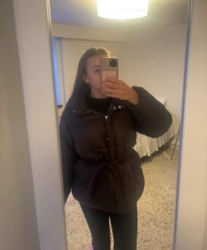 Brun pufferjacka med bälte - Säljer en brun pufferjacka med bälte i midjan och hög krage. Jackan har dragkedja och knappar framtill, samt långa ärmar. Man kan ta bort bandet. Köpt i slutet av 2025 och knappt använd
