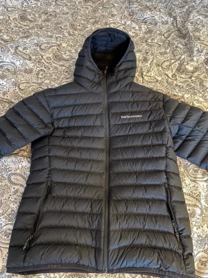 Svart dunjacka Peak Performance - Svart dunjacka från Peak Performance med huva och dragkedja framtill. Jackan har ett quiltat mönster och två sidofickor med dragkedja. Perfekt för kalla dagar och har en clean, sportig look som funkar till det mesta.