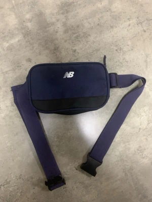 Mörkblå midjeväska från New Balance - Säljer en mörkblå midjeväska från New Balance med justerbart band och praktisk dragkedja. Väskan har en stilren design med svart detalj på framsidan och logga i vitt. Perfekt för att bära med sig det viktigaste och har ett fack med dragkedja inuti.