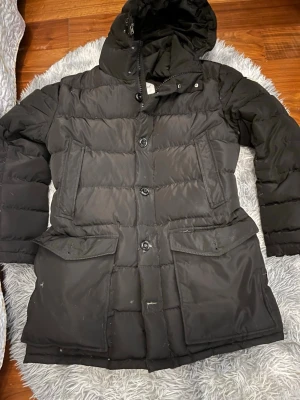 Svart Moncler dunjacka med huva - Säljer en svart dunjacka från Moncler med huva och stora fickor framtill. Jackan har både dragkedja och knappar, samt det klassiska Moncler-logotypen på ärmen. Perfekt för kalla dagar och riktigt snygg till vinterlooken.