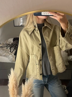 Grön jeansjacka med broderade detaljer - Säljer en grön  jeansjacka med snygga broderade hålmönster längs nederkanten och fickorna. Jackan har en loose passform, stora fickor framtill och knappar. Perfekt för dig som gillar unika detaljer och chill stil.