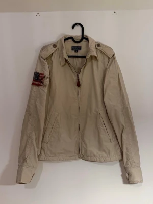 Beige militärjacka från Polo Ralph Lauren Jeans Company - Snygg beige vindjacka från Polo Jeans Company med dragkedja och cool amerikansk flaggpatch på ärmen. Jackan har krage, knappdetaljer på axlarna och fickor fram. Perfekt för dig som gillar klassisk streetstil och vill ha något som sticker ut.