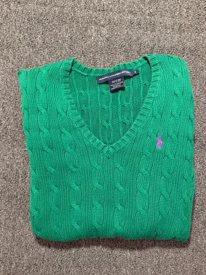 Grön stickad tröja från Ralph Lauren - Snygg grön kabelstickad tröja från Ralph Lauren Sport med lila broderad logga på bröstet. Tröjan har v-ringad hals, långa ärmar och ribbade muddar. Tillverkad i 100% bomull, perfekt för dig som gillar klassisk stil med en färgklick.