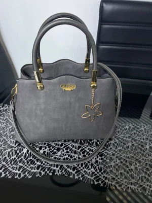 Grå handväska från Guess - Snygg grå handväska från Guess med guldfärgade detaljer och en dekorativ blomma i metall. Väskan har både handtag och axelrem, vilket gör den enkel att bära. Stilren design med mjukt material och elegant logga framtill.