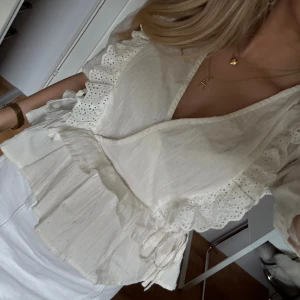 Vit omlottblus med volang och spets - Superfin vit omlottblus med volangdetaljer och spets längs kanterna. Blusen har korta ärmar, knyt i midjan och v-ringning. Perfekt för en fräsch och somrig look. Materialet känns lätt och luftigt, vilket gör den skön att bära under varma dagar.