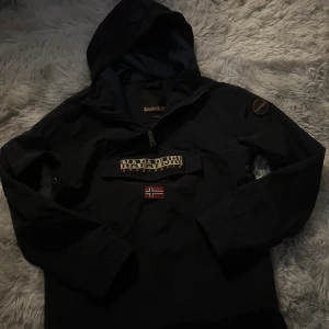 Svart vindjacka från Napapijri - Svart vindjacka från Napapijri med huva och halv dragkedja. Väldigt fint skick och är i storlek xs. Ny pris:1400kr