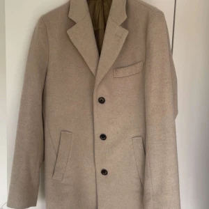 John Henric rock  - Stilren beige kavaj med tre knappar framtill och klassisk krage. Kavajen har två snedställda fickor och en bröstficka. Materialet är mjukt och känns lyxigt, perfekt för dig som gillar en clean och tidlös look.