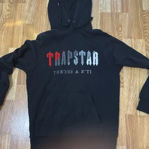 Svart Trapstar hoodie med brodyr - Svart hoodie från Trapstar med broderad logga i rött, grått och vitt på bröstet. Klassisk känguruficka framtill, huva med snörning och mjukt material. Perfekt för dig som gillar streetwear och vill sticka ut med en fet logga.