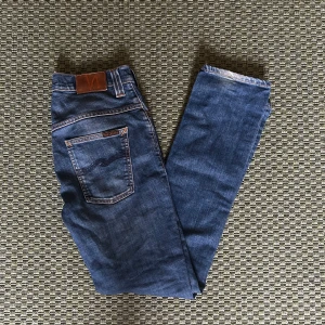 Blå jeans från Nudie Jeans - Säljer ett par klassiska blå jeans från Nudie Jeans med snygga kontrastsömmar och ikonisk broderad bakficka. Jeansen har normal passform och raka ben, tillverkade i slitstarkt denimtyg. Perfekta för dig som gillar stilrena och tidlösa jeans.