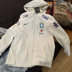 Vit Nike Brasil regnjacka med huva - Vit regnjacka från Nike med Brasil-logga och sponsormärken. Jackan har huva med snörning, dragkedja framtill och långa ärmar. Perfekt för fotbollsfans som vill hålla sig torra och snygga. Snygga detaljer i grönt och lila tryck.