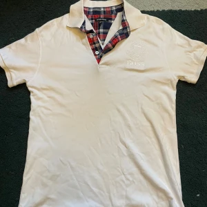 Vit pikétröja från Hugo Boss M - Vit pikétröja från Hugo Boss med broderad logga på bröstet och snygg rutig detalj på insidan av kragen i rött, blått och vitt. Klassisk pikékrage och korta ärmar. Perfekt för en clean och stilren look.