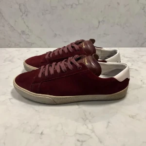 Vinröda Saint Laurent sneakers i mocka - Snygga vinröda sneakers från Saint Laurent i mocka med vita detaljer på hälen och klassisk snörning. Skorna har en beige sula och Saint Laurent-logga i guld på skon. Perfekta för dig som vill ha en stilren och lyxig look. Stlr 41 men passar 42. Priset är inte hugget i sten så skicka prisförslag.