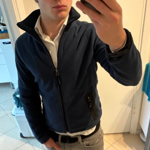 Mörkblå fleecejacka Key West XS - Mörkblå fleecejacka från Key West med hög krage och hel dragkedja. Jackan har två svarta fickor med dragkedja framtill och logodetaljer på ärm och nacke. Perfekt för kyliga dagar. Bra skick, men armspännena har lossnat (cardborren)