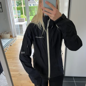 Svart Gore-Tex vindjacka från Peak Performance - Svart vindjacka från Peak Performance med vit dragkedja och ventilerande zip under armarna. Jackan är tillverkad i Gore-Tex Paclite Shell, har hög krage och praktiska fickor med dragkedja. 