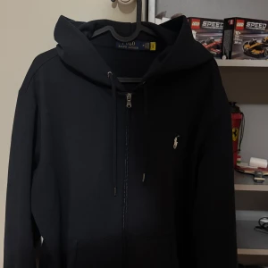 Marinblå hoodie från Polo Ralph Lauren - Marinblå hoodie från Polo Ralph Lauren med dragkedja framtill, snörning i huvan och klassisk broderad logga på bröstet. Tröjan har känguruficka och är långärmad, perfekt för en chill och stilren look.