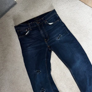 Mörkblå skinny jeans från Nudie - Snygga mörkblå skinny jeans från Nudie med slitna detaljer på benen och klassisk femficksdesign. Jeansen har en smal passform och är gjorda i mjukt denimtyg med stretch för extra komfort. Perfekta för en avslappnad och trendig look. Jeansen är lite slitna vilket ger dem en snygg look! Dom är i storlek 29/30