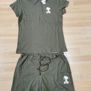 Olivgröna Snoopy pyjamas i 2 delar  - Olivgröna pyjamas från Snoopy i 100% bomull med snörning och knappar framtill. Shortsens ena ben har en stor ficka med ett tryck av Snoopy. Perfekta för chill dagar och sommarhäng.
