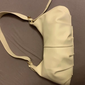 Beige axelväska i skinnimitation - Stilren beige axelväska med bred axelrem och snygga veckade detaljer. Väskan har en mjuk, slät yta och guldfärgade metalldetaljer vid remmarna. Perfekt för dig som gillar minimalistisk och trendig design.