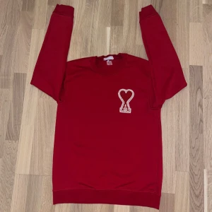 Röd sweatshirt från Ami Paris - Röd sweatshirt från Ami Paris med ikoniskt hjärta och A-logga broderad på bröstet. Klassisk rund hals och långa ärmar. Tillverkad i mjuk bomull för en skön och avslappnad vibe.