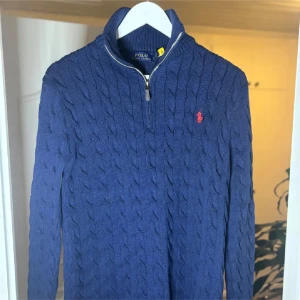 Ralph Lauren half zip marinblå  - Kabelstickad marinblå tröja från Polo Ralph Lauren med halv dragkedja och röd broderad logga på bröstet. Tröjan har hög krage och är långärmad, perfekt för kyliga dagar. Klassisk och stilren design som funkar till allt.