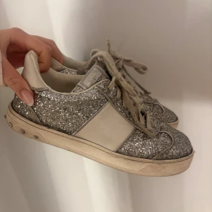 Valentino Garavani glittriga sneakers - Snygga sneakers från Valentino Garavani med silverglittriga paneler, vita detaljer och coola nitar runt sulan. Skorna har snörning och en platt sula. Perfekta för dig som vill sticka ut med lite extra bling i vardagen. Slitna därav priset 