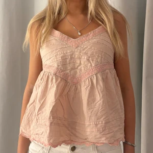 Ljusrosa blus med spetsdetaljer - Supersöt ljusrosa blus med tunna axelband och v-ringad hals. Blusen har broderade spetsdetaljer i rosa längs halsringningen och midjan, samt vågig spetskant nedtill. Luftig och flowy passform, perfekt för varma dagar.