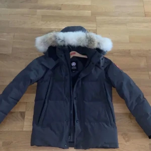 Svart dunjacka från Canada Goosee - Säljer en svart dunjacka från Canada Gooseee med avtagbar huva och pälsdetalj. Jackan har lång ärm, dragkedja och klassisk patch på ärmen. Perfekt för kalla vinterdagar och har en snygg, stilren look.