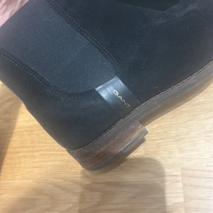 Svarta chelsea boots från Gant i mocka - Snygga svarta chelsea boots från Gant i mjuk mocka med klassisk rund tå och låg klack. Skorna har elastiska paneler på sidorna och en stilren design som passar till det mesta. Perfekta för dig som gillar en clean och tidlös look.