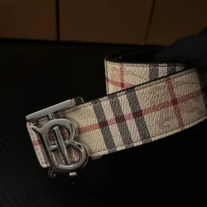 Burberry bälte med TB-spänne - Säljer ett Burberry bälte med ikoniskt TB-spänne i silverfärgad metall. Finns i klassiskt rutig beige med svarta och röda detaljer samt i svart med diskret rutig struktur och präglad riddare. Båda är tillverkade i exklusivt skinn och har en lyxig känsla.