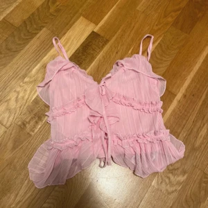 Rosa volangig blus från Amisu storlek  XS - Supersöt ljusrosa blus från Amisu i storlek XS. Toppen har tunna axelband, volanger och snörning framtill. Perfekt för dig som gillar gulliga och trendiga plagg! Toppen är helt oanvänd med prislappen kvar därav priset. 