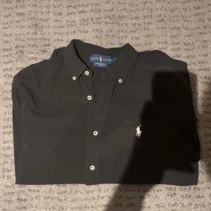 Svart skjorta från Ralph Lauren storlek M - Svart skjorta från Ralph Lauren i storlek M, tillverkad i lätt mesh-bomull. Klassisk design med knappar framtill och vit broderad logga på bröstet. Skjortan har lång ärm och en normal passform, perfekt för dig som gillar stilrena plagg med ikonisk touch. Jag på bilden är 186 och väger 71
