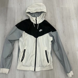 Nike vindjacka med huva i svart och grått - Nike vindjacka med huva i svart, grå och vit. Jackan har dragkedja framtill, två fickor med dragkedja och meshfoder. Snygg sportig design med logga på bröstet och snörning i huvan. Perfekt för dig som gillar streetwear och sportig stil.
