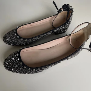 Svarta ballerina med nitar och strass - Säljer ett par svarta ballerina med rund tå, dekorerade med silvriga nitar och strass över hela skon. Skorna har en låg klack och ett smalt ankelband med spänne. 