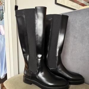 Svarta höga stövlar - Snygga svarta stövlar från H&M i storlek 40. De har en stilren design med rund tå och platt sula, tillverkade i syntetmaterial och textil. Stövlarna är höga och har elastiska paneler på sidorna för extra komfort och passform.