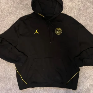 Svart PSG x Jordan hoodie med gula detaljer - Svart hoodie från Jordan x PSG med gula detaljer och tryck. Stor Paris Saint-Germain-logga på ryggen, gul Jumpman-logga och PSG-märke på bröstet. Huva med snörning och känguruficka framtill. Perfekt för dig som gillar streetwear och fotboll.