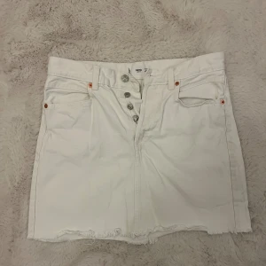 Vit jeanskjol från Mango - Säljer en vit jeanskjol från Mango med rå kant nedtill och silverfärgade knappar framtill. Kjolen har klassiska fickor både fram och bak samt hällor för bälte. Perfekt för dig som gillar en clean och fräsch look.