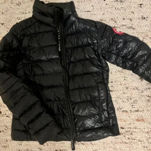 Svart dunjacka från Canada Goose - Säljer en svart dunjacka från Canada Goose med klassisk logga på ärmen. Jackan har quiltad design, hög krage och dragkedja framtill. Materialet är glansigt och lätt, perfekt för kalla dagar. Snygg och stilren modell som funkar till många olika outfits.