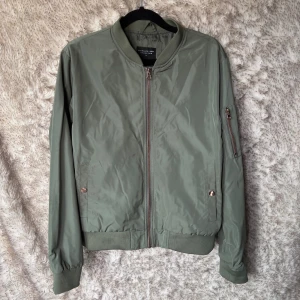 Olivgrön bomberjacka från The Black Label - Snygg olivgrön bomberjacka från The Black Label Collection i storlek M. Jackan har ribbade muddar, dragkedja i kopparfärg och en extra ficka med dragkedja på ärmen. Tillverkad i polyester och har en klassisk bomberpassform.