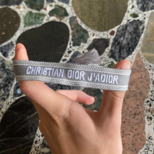 Grå Christian Dior J'Adior armband - Snyggt grått armband från Christian Dior med broderad text 'J'Adior' i vitt. Armbandet har två dekorativa tofsar och är gjort i ett mjukt tygmaterial. Perfekt accessoar för att lyfta din outfit med lite lyxig vibe.