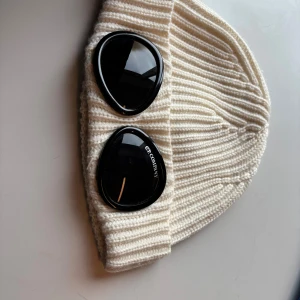 Vit ribbad mössa C.P. Company - Säljer en vit ribbstickad mössa från C.P. Company med två svarta glasögon-linser på uppviket. Mössan är tillverkad i mjuk ull och har en unik, urban look som sticker ut. Perfekt för dig som vill ha något annorlunda och snyggt på huvudet.