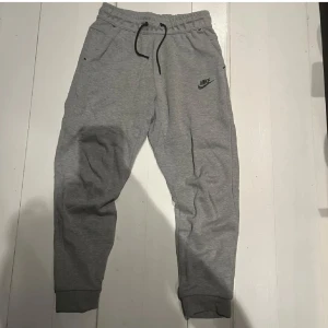 Grå mjukisbyxor från Nike - Grå mjukisbyxor från Nike med svart logga på vänster ben. Byxorna har resår och snörning i midjan, muddar vid bensluten och praktiska fickor, varav en bakficka med dragkedja. Perfekta för chill eller träning.