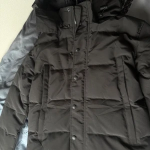  Canada Goose - Säljer en svart Canada Goose jacka med klassisk logga på ärmen. Jackan har huva, dragkedja och tryckknappar framtill samt två stora fickor. Perfekt för kalla dagar och riktigt snygg streetstil. Påsen och pälsen följer med. 